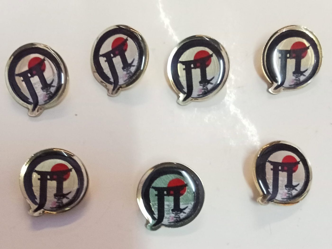 Round gold-rimmed lapel pins with a red sun and torii gate logo under a glossy dome finish